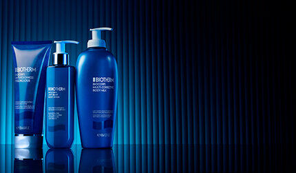 Three Biotherm dark blue bottles of body care range, set against a textured vertical striped background with dramatic lighting.